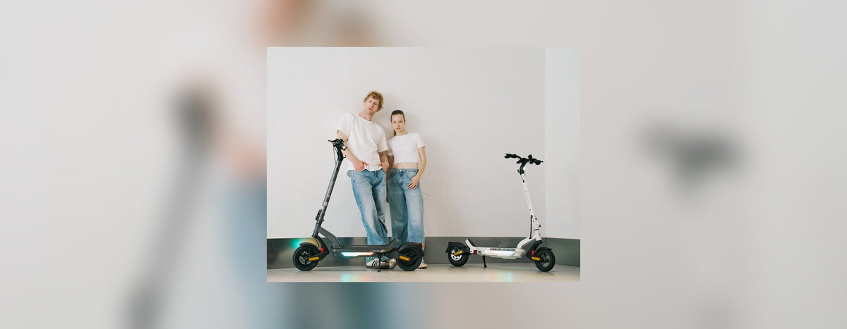 Two people standing with a black and a white isinwheel electric scooter against a plain studio wall