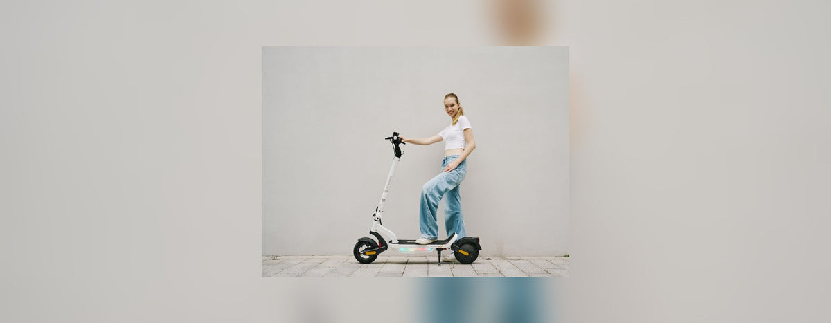 Woman standing with a white isinwheel electric scooter in front of a plain pale wall