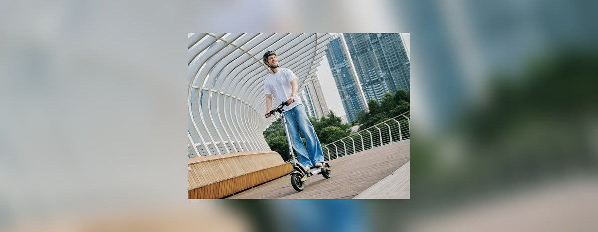 Man riding a black isinwheel electric scooter across a curved footbridge with city tower blocks behind