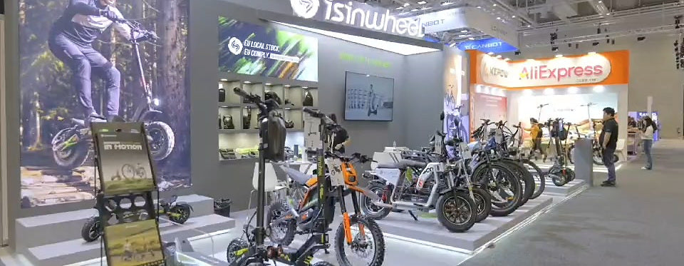 The isinwheel stand at IFA Berlin 2026: backlit logo wall, an accessory wall, screens, and a display plinth of electric scooters and fat-tyre e-bikes