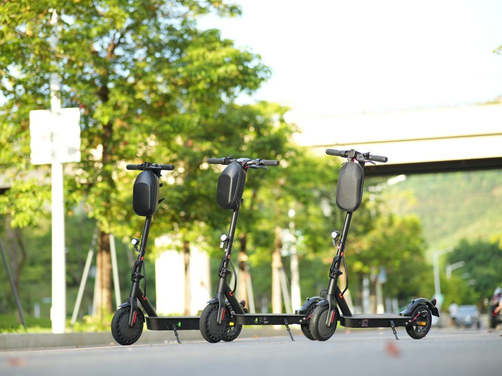Three early isinwheel commuter scooters with handlebar bags, parked by a road