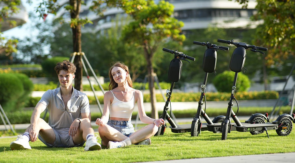 Two young people sitting on the grass in a park, with three isinwheel electric scooters parked behind them